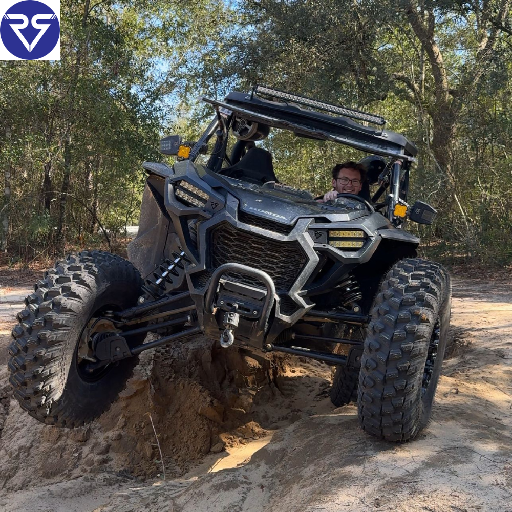 Black Honda UTV with off-road tires climbing uneven dirt trail, featuring LED light bar and winch, driven through wooded terrain by a rider.