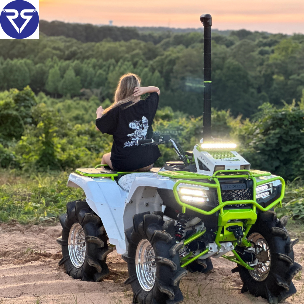 Rider sitting on modified Honda ATV with mud tires and LED lights, overlooking scenic forest view at sunset, ready for off-road adventure.
