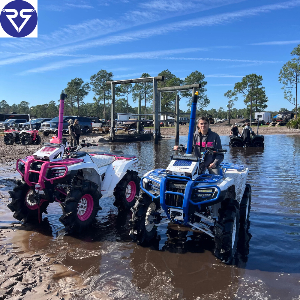 Two modified Honda ATVs with snorkels and mud tires parked in shallow water, riders preparing for off-road riding at a muddy trail park.
