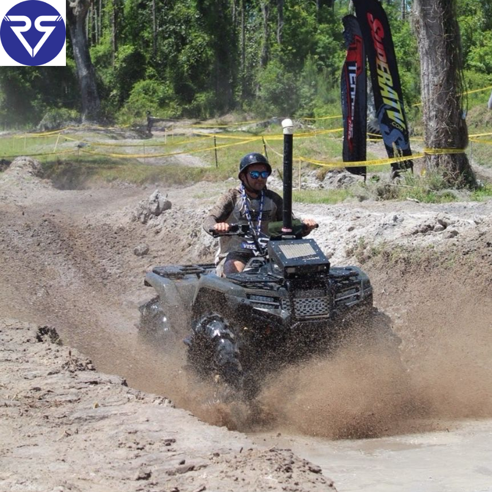 Rider wearing helmet drives an ATV through muddy trail, splashing water and dirt, surrounded by trees and off-road course markers.