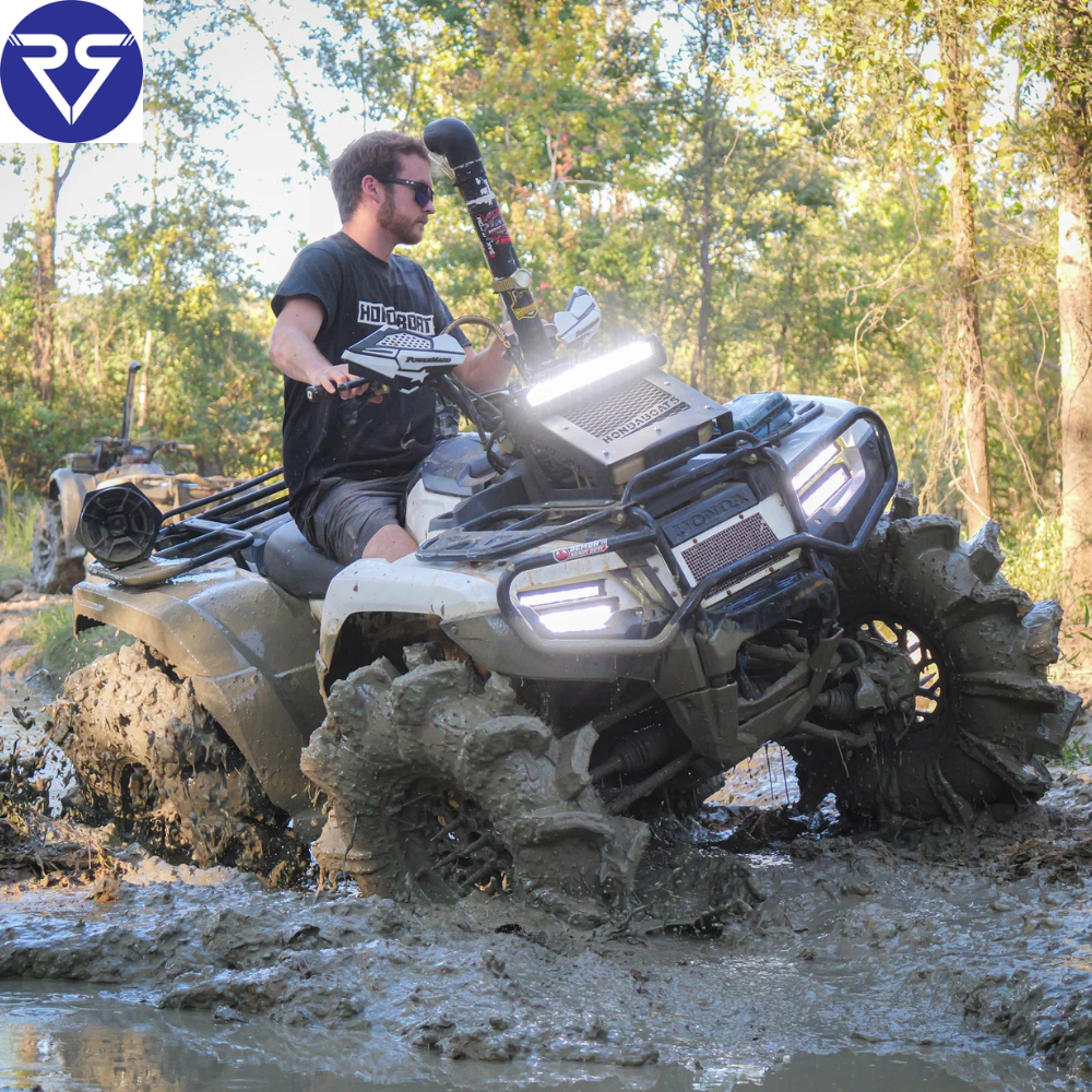 Rider on modified Honda ATV driving through deep mud, equipped with snorkel, LED light bar, and aggressive tires on a rugged forest trail.