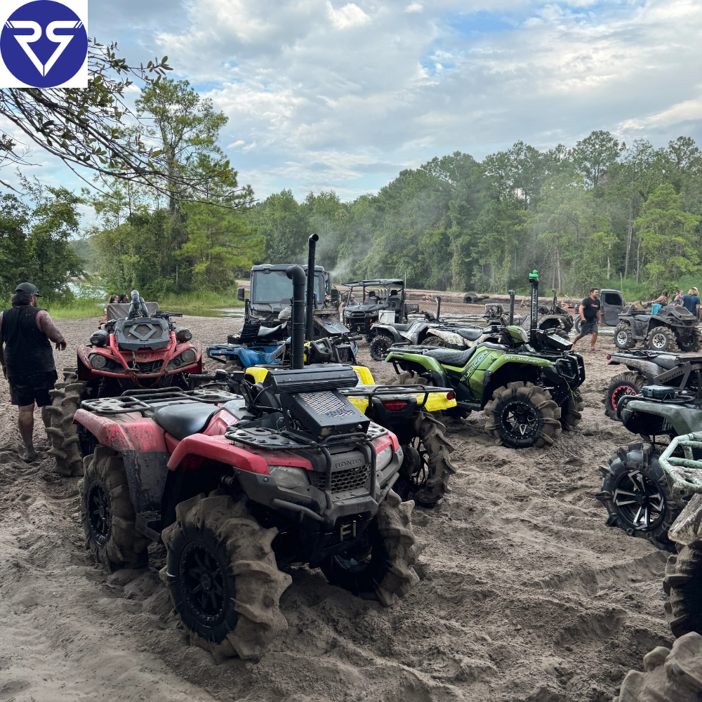 Group of modified ATVs with mud tires gathered on sandy trail, riders preparing for off-road adventure near forest under cloudy sky.