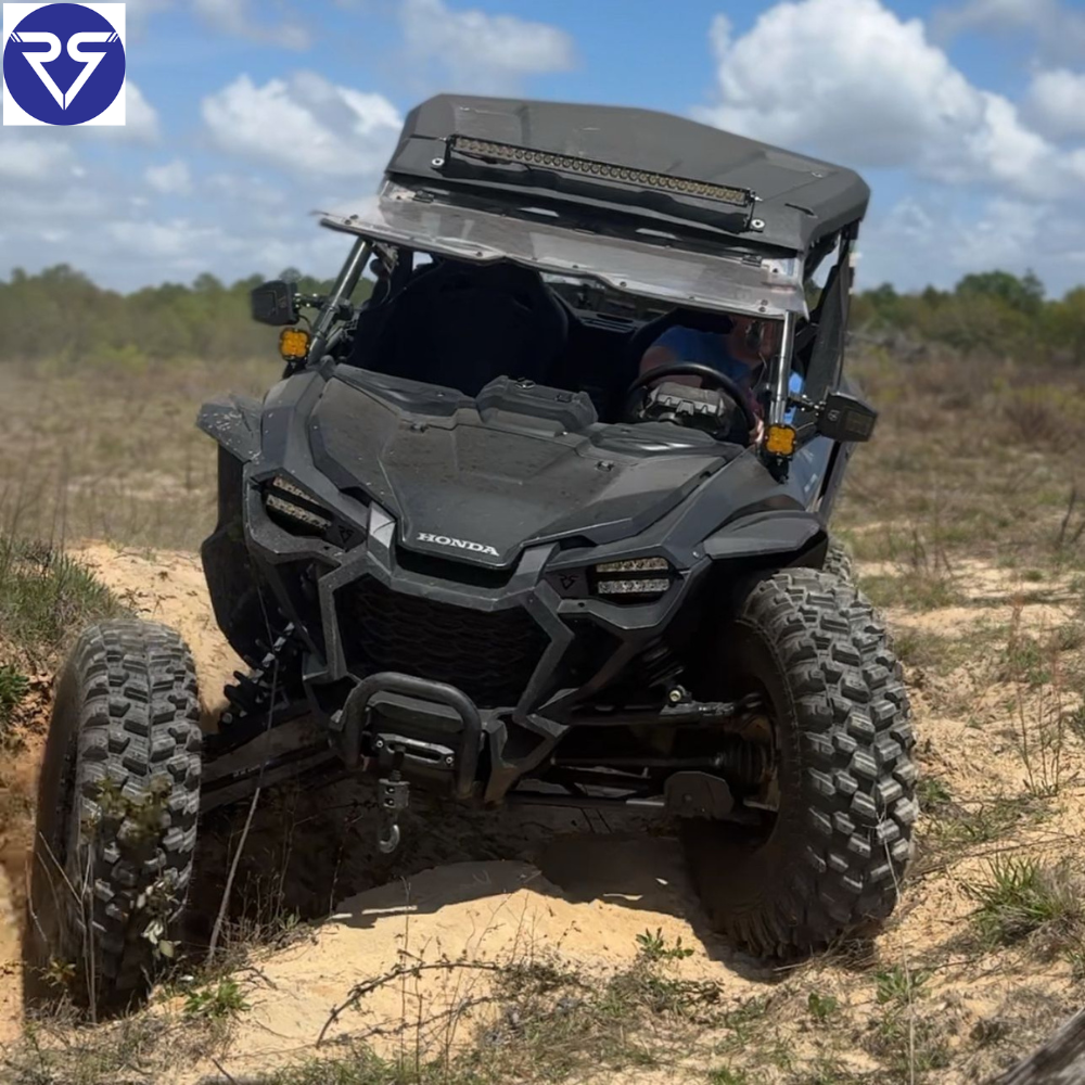 Black Honda UTV with aggressive off-road tires driving over sandy terrain, equipped with LED light bar and rugged suspension under clear sky.