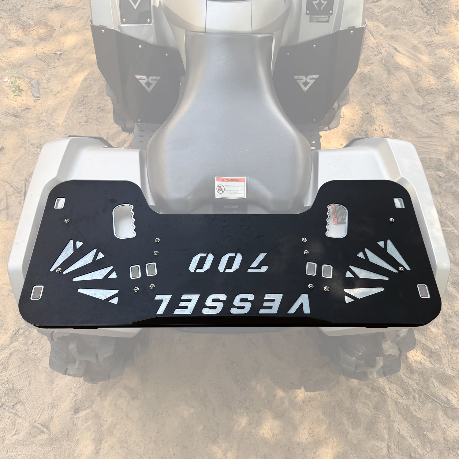 A black metal ATV rear bumper plate with silver 'VESSEL 700' lettering and geometric cutouts, shown mounted on the back of a white and gray ATV on a sandy ground.