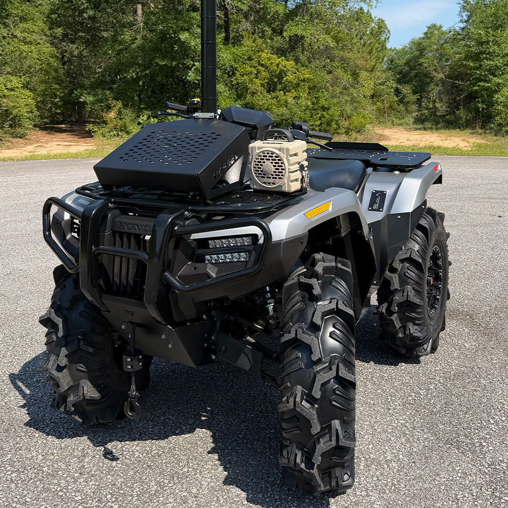 A black and silver Polaris ATV parked on an asphalt surface with trees in the background. The ATV is equipped with a mounted black storage box, a beige speaker, and a vertical antenna.